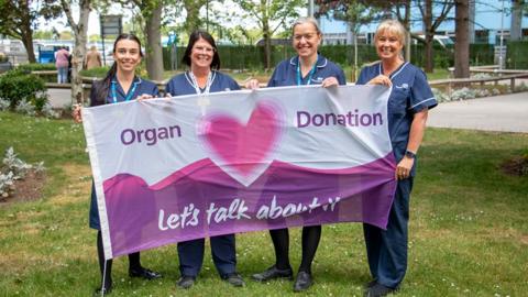 Four organ donation nurses in blue uniforms hold the flag outside of the Hull Royal Infirmary. The flag is purple and white and has a heart in the middle with the words Organ Donation Let's Talk About It.