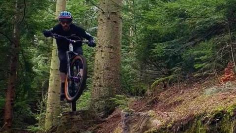 A mountain biker is flying mid-air while he completes a trick. The photograph was taken in Coombs Wood, Armathwaite.