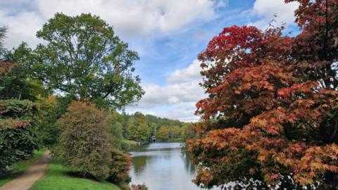 green and red trees either side of a lake with a path on the left, under a cloudy, blue sky