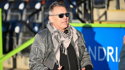 Dale Vince walking at Forest Green Rovers' Bolt New Lawn Stadium. He is wearing a grey coat, white, black and grey patterned scarf and black sunglasses