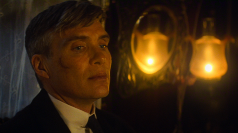 Cillian Murphy, playing Tommy Shelby in a film still, looking away from the camera. He has a scar on his face and lights glowing yellow behind him. His hair is slightly grey. 