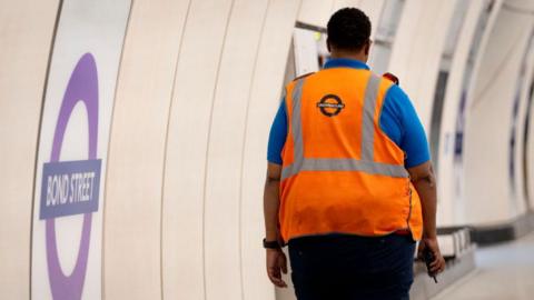 A TfL worker walks along the Elizabeth Line platform at Bond Street with a walkie-talkie in hand and a high-visibility jacket. He faces away from the camera.