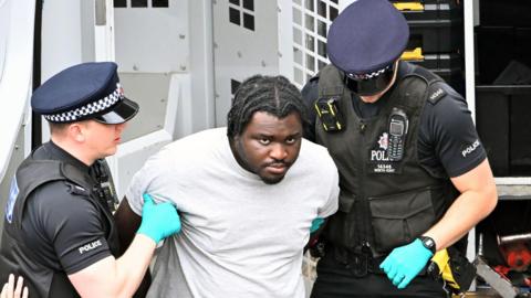 Anthony Esan is seen being held by two police officers, who are wearing uniforms and gloves. Esan is wearing a grey top and has his hands behind his back.
