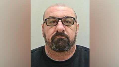 Christopher O'Neill's head and shoulders mugshot. He has shaved hair, black-rimmed glasses and a black beard. He is wearing a black t-shirt and is in front of a grey background