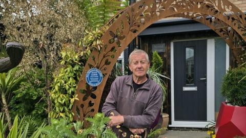 Steve Damon is pictured in his front garden. He is standing under an ornate rusted metal archway on which there is a blue plaque which reads 'Steve Damon, gold award, best front garden, East Midlands in Bloom 2025'. The black front door of Steve's house can be seen just beyond the arch. The garden is filled with tropical plants and sculptures.