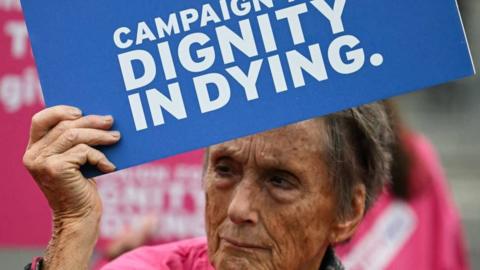 A woman holds up a sign that says: Campaign for dignity in dying. The sign is blue and the woman has grey hair.