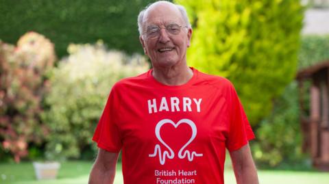 Harry Newton, 88, who has a bald head with white patches of hair on the sides and round-framed glasses, smiles at the camera in front of a garden lawn. He is wearing a red t-shirt with the logo of the British Heart Foundation in white, and the word 'Harry'. 