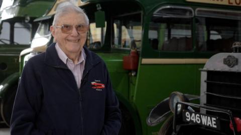 A man with glasses and a dark blue cardigan. He is smiling. He is standing in front of a row of vintage green buses.