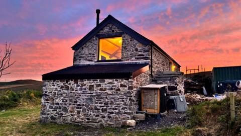 A stone cosy-looking cottage in front of pink skies at sunset, with materials from the renovation project on the right hand side of the building.