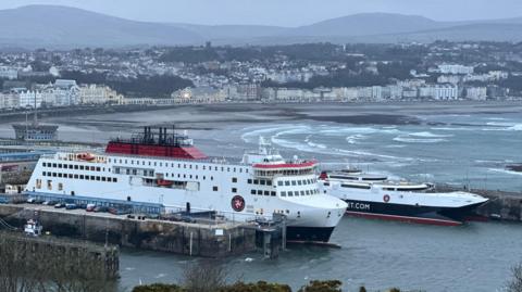 The Manxman and Manannan ferries, which are both white, red and black, moored in Douglas Harbour. Choppy seas in Douglas Bay are in the background.