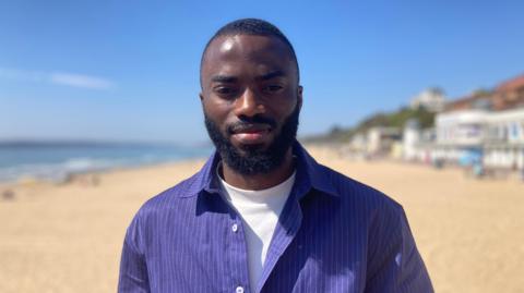 A man stands on a beach, wearing a striped blue shirt with a white t-shirt underneath. The sun is shining, the sea is blue and the sand is yellow. You can see some beach huts or businesses in the background in the far right hand corner
