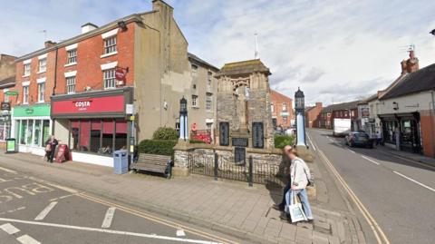 Streetview image of an end of terrace building, with a traditional memorial garden separating the shop from a road junction