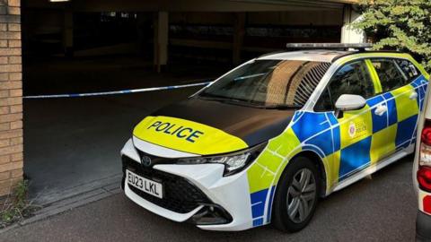 A police car parked in front of a taped-off car park.