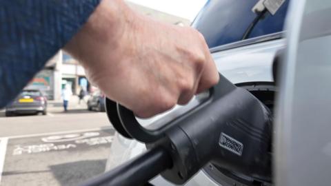 A close up image of a hand holding an electric car charger as it plugs into the socket on a silver car.  