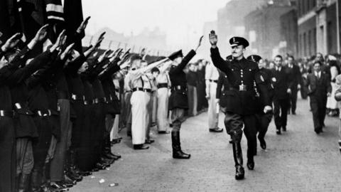 A man walks in uniform with his arm stuck out in the air. People line the streets with their arms out in the air also. 