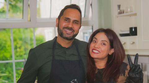 A man and a woman with their arms round each other, smiling at the camera. The woman is making a peace sign with her hand.
