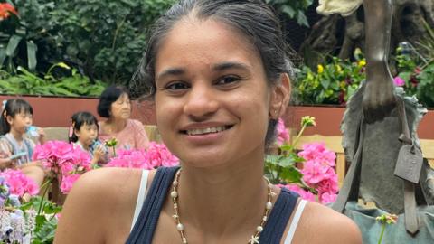 Elina Patel smiles at the camera wearing a blue T shirt and a necklace with ornamental gardens in the background