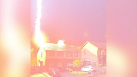 A lightning bolt is seen striking a detached house. The image is tinged with pink and orange capturing the moment the lightning hits the roof of the house. It is in a residential area with trees, cars and another house on the left hand side of the building.