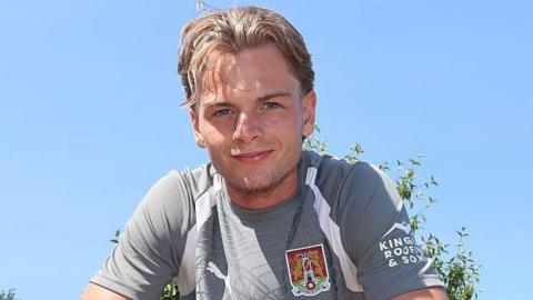 A smiling Logan Briggs wearing a grey Northampton Town training top faces the camera