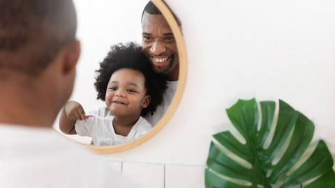 A boy grins at his father in the mirror while brushing his teeth.