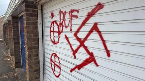 A close-up of a white garage door with some red graffiti of a swastika, "BUF" and two circles with crosses in them.