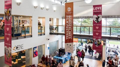 The inside of a theatre building lobby with two floors. The entrance wall has glass double doors and glass floor to ceiling windows. There are people walking around on the lower level near the entrance. There is a seating area inside and outside. On the floor above is a walkway stretching across the space with seating on one side. There are banners hanging from the ceiling that say "borderlines film festival" and "love malvern theatres"