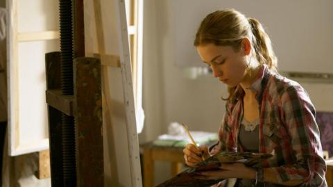 A fine art student painting in a studio. She is mixing paints on a palette. A white canvas on an easel stands in front of her. She has mousey blonde hair and is wearing a chequered shirt.