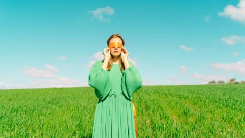 Woman in a green dress stands in a field wearing a blind fold