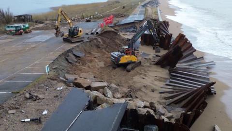 Two yellow diggers work on repairs to the A379 in Devon after storm damage to sea defences washed away parts of the road. The road is next to a beach and the sea. Waves are washing in from the sea. Large pieces of metal from the sea defences are scattered along the sandy beach.