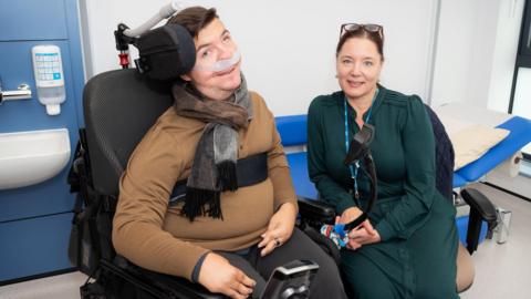 A man in a wheelchair that has a small screen attached to it. There is a pad on one side of his head which he is leaning against, and he has a ventilator going into his nose. He is wearing a brown long-sleeved top. Next to him is a woman wearing a forest green dress with a blue lanyard around her neck. Her hair is tied back and she has glasses on her head.