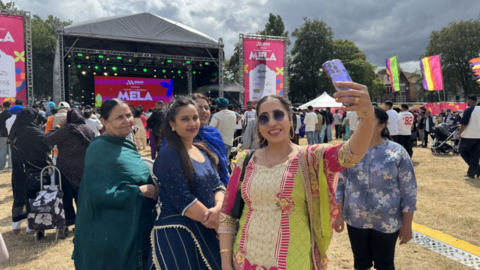 A woman takes a selfie with a group of other women, with crowds of people standing behind them, looking at a stage.
