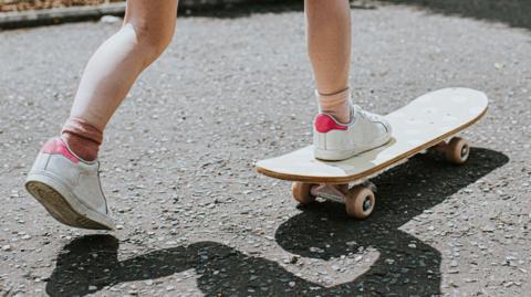 A stock image of a child on a skateboard. The child is wearing white and pink trainers and mismatched pink socks. 
