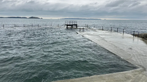 A diving board on the edge of an open air seawater bathing pool on a grey-weather day. Both the sea and pool are at the level of the edge of the pool, and there are islands in the background.