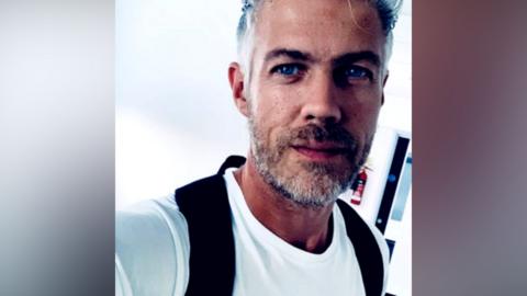 A man with a greying beard is looking at the camera and appears to be taking a selfie. He is wearing a white t-shirt and a black rucksack.