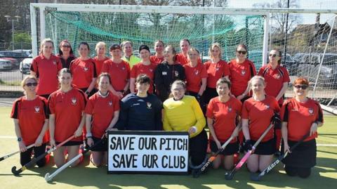 Southwick's women's team lined up in the goal mouth of their pitch with their hockey sticks and red tops with a sign that reads "save our pitch, save our club".