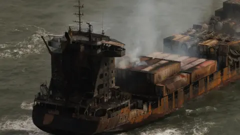 A rusted and burnt Solong cargo ship with smoke coming from the top of it in waves of the sea