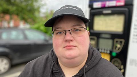 Ellie Walker, wearing a baseball cap and hoodie, in front of a parking machine