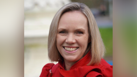 A woman with bobbed blonde hair wears a red jacket and smiles at the camera.
