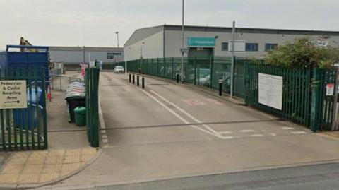 Entrance to Hazel Court tip on James Street in York. There are green railings lining the entrance road, with a sign to the left pointing to a 'Pedestrian and Cyclist Recycling Area'. To the right behind the railings there's a large industrial building, with 'Be Independent' above the doors.