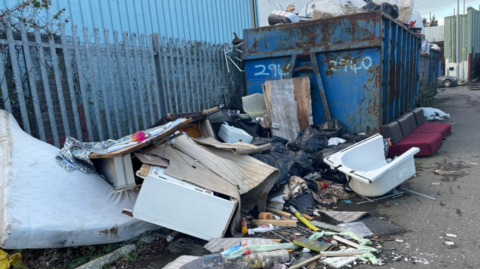 An image of rubbish, including a bath, spread out on a road. A huge, blue metal container is in the background next to a fence.