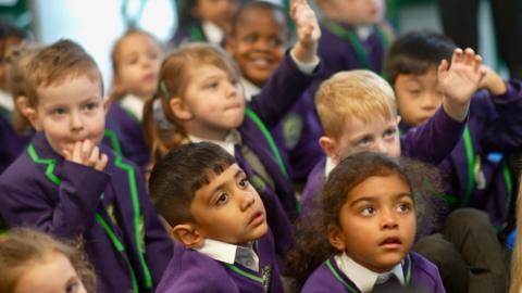 Children at Chapel Hill Primary School looking to the side of the camera. Some have their hands up. Their uniform is purple and green.