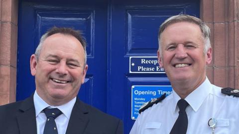 David Allen and Darren Martland stand side by side outside a blue police station door. They are both smiling brightly at the camera. Allen has short dark/grey hair and is wearing a dark blue suit and tie. Martland is in a police uniform, which consists of a white shirt, black epaulets and an identification card clipped to his pocket. He also has short greying hair.