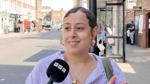 Person speaking into a BBC microphone on a sunny urban street with shops and a bus stop in the background.