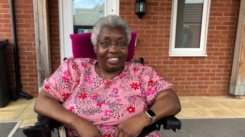 A picture of a lady sat in a wheelchair smiling at the camera. She has grey hair and is wearing glasses. She is wearing a watch on her left wrist. She is sat in front of a brick house.
