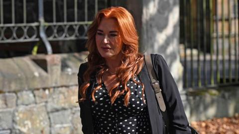 Anne Donaghy, a woman with red hair, wearing a dark jacket, and black blouse with white spots.