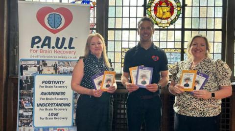 Three people holding a purple and a yellow leaflet stood in front of a stained glass window inside a building. They are stood next to a banner with multiple images and reads 'P.A.U.L For Brain Recovery'. The woman on the left is wearing a black spotty blouse and black trousers. She has long blonde hair. The man is wearing a black polo shirt with a red and blue logo on and black trousers. He has short brown hair. The woman on the right is wearing a black and white patterned blouse and black trousers. She has light-coloured hair tied back. They are all smiling at the camera. 