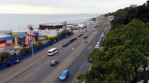 A stock shot of Southend seafront, taken from an elevated position. It shows a four-lane road running alongside the promenade. There are cars driving in both directions. The sea is on the right. Tourist attractions including a helter skelter are also on the right of the picture. There are trees on the left.