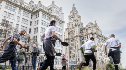 Shot of several chefs in their chef whites running round the church gardens with pancake and pan in hand. The Liver Building can be seen in the distance.