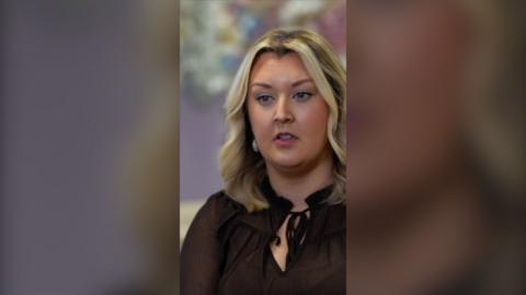 A woman with wavy brown hair with highlights looks off camera. She is wearing a black shirt tied at the neck.