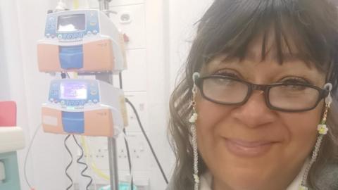 Rhian Gordon, wearing brown framed glasses, smiles at the camera as she receives chemotherapy in hospital. 